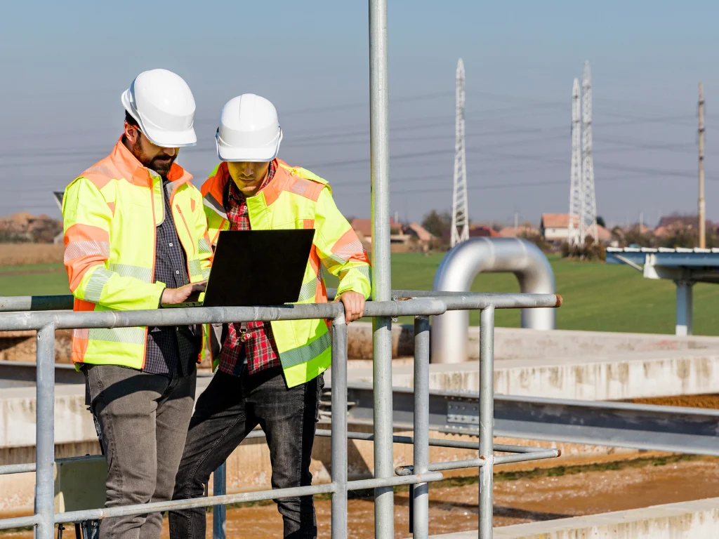 Two engineers in high-visibility jackets and hard hats reviewing laptop at industrial site