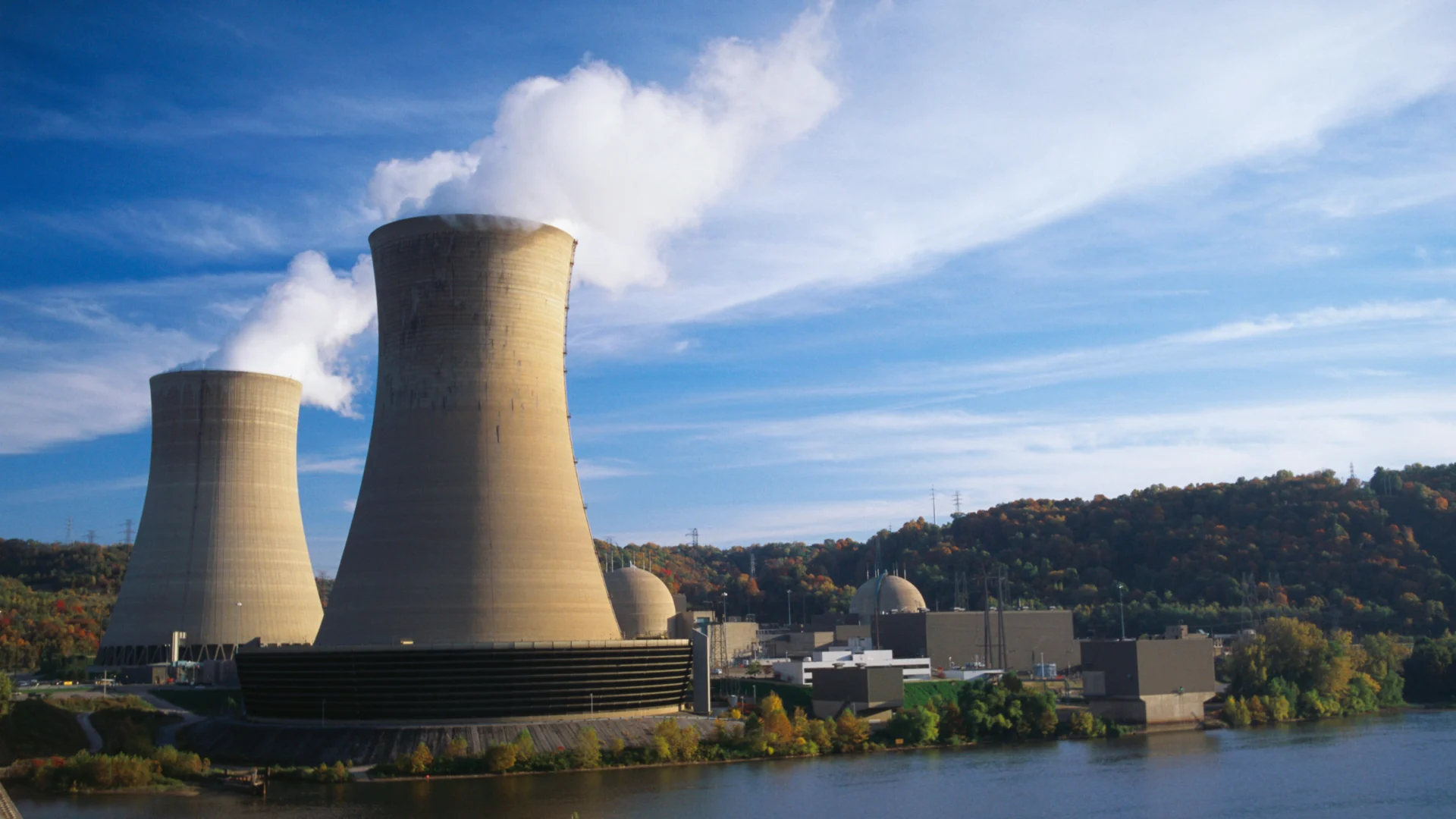 Cooling towers emitting steam at a power plant beside a river, with industrial buildings and wooded hills in background.