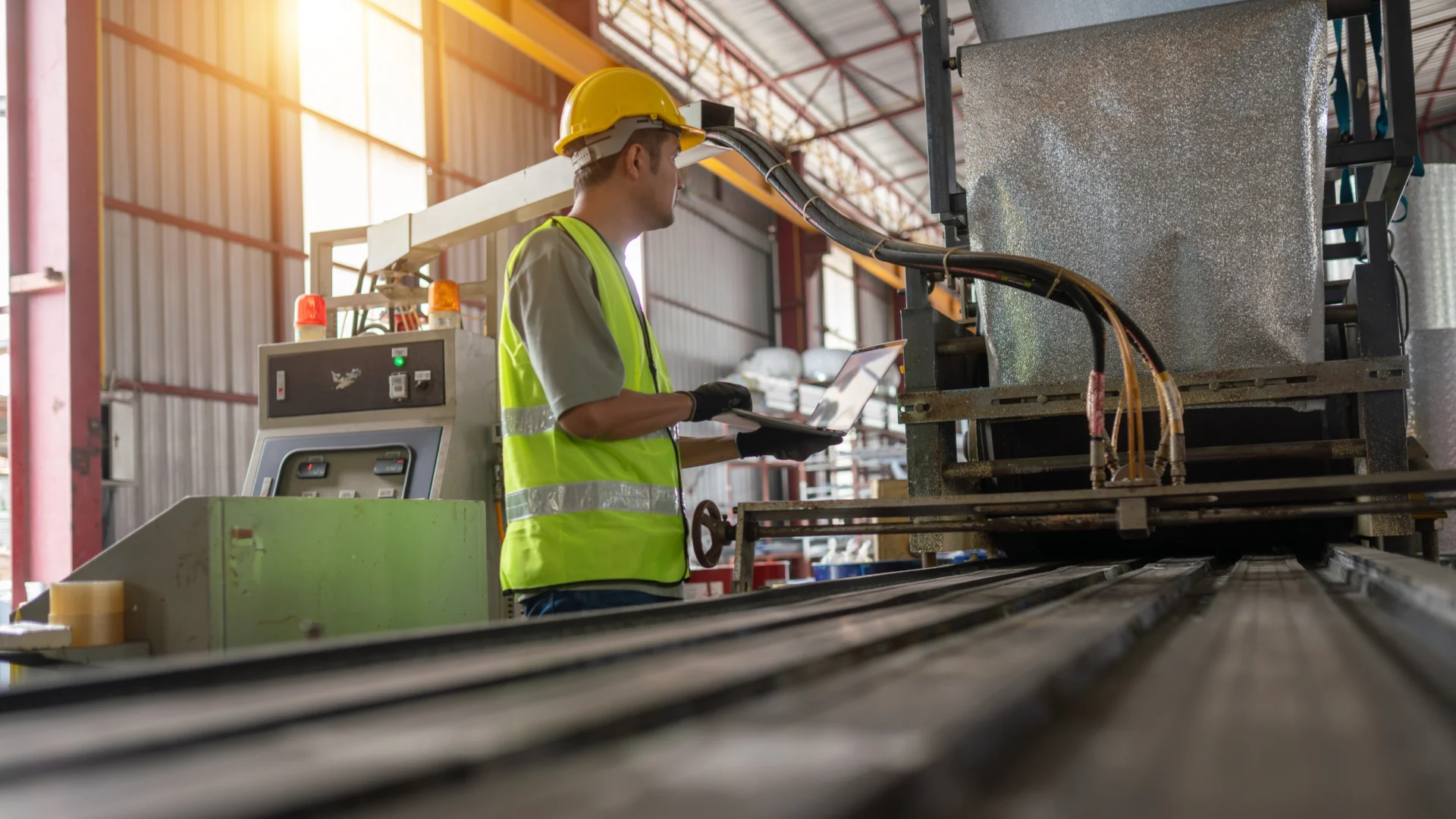 Technician in safety gear using laptop to monitor industrial machinery on production line inside manufacturing facility.