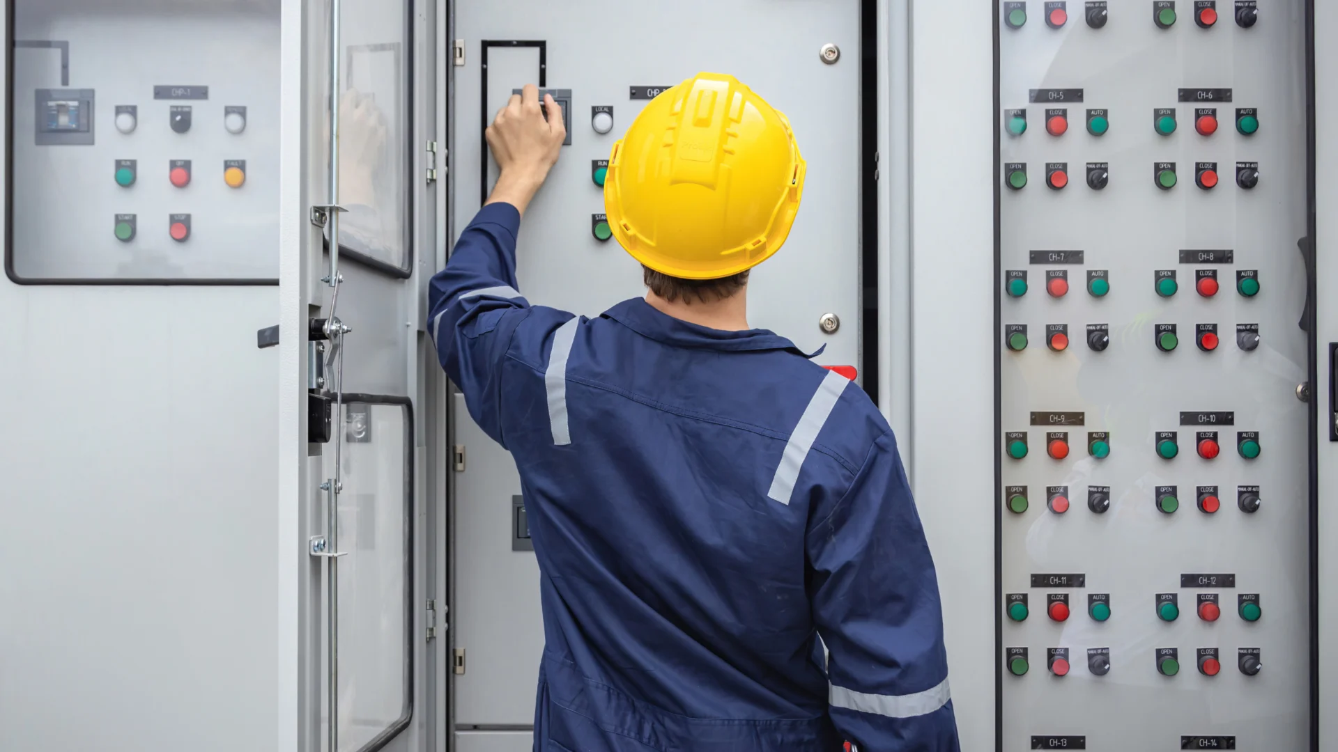 Technician in safety helmet operating electrical control panel with switches and indicators inside industrial cabinet.