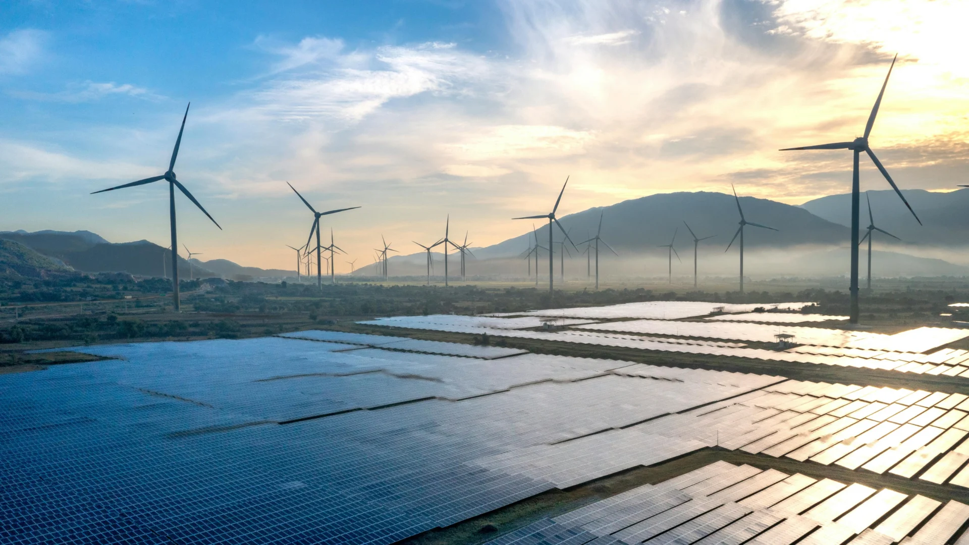 Solar panels and wind turbines backed by a sunset and foothills.